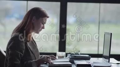 商业上的问题，年轻的女企业家坐在靠窗的桌子旁，紧张地一边远程工作一边打着笔记本电脑